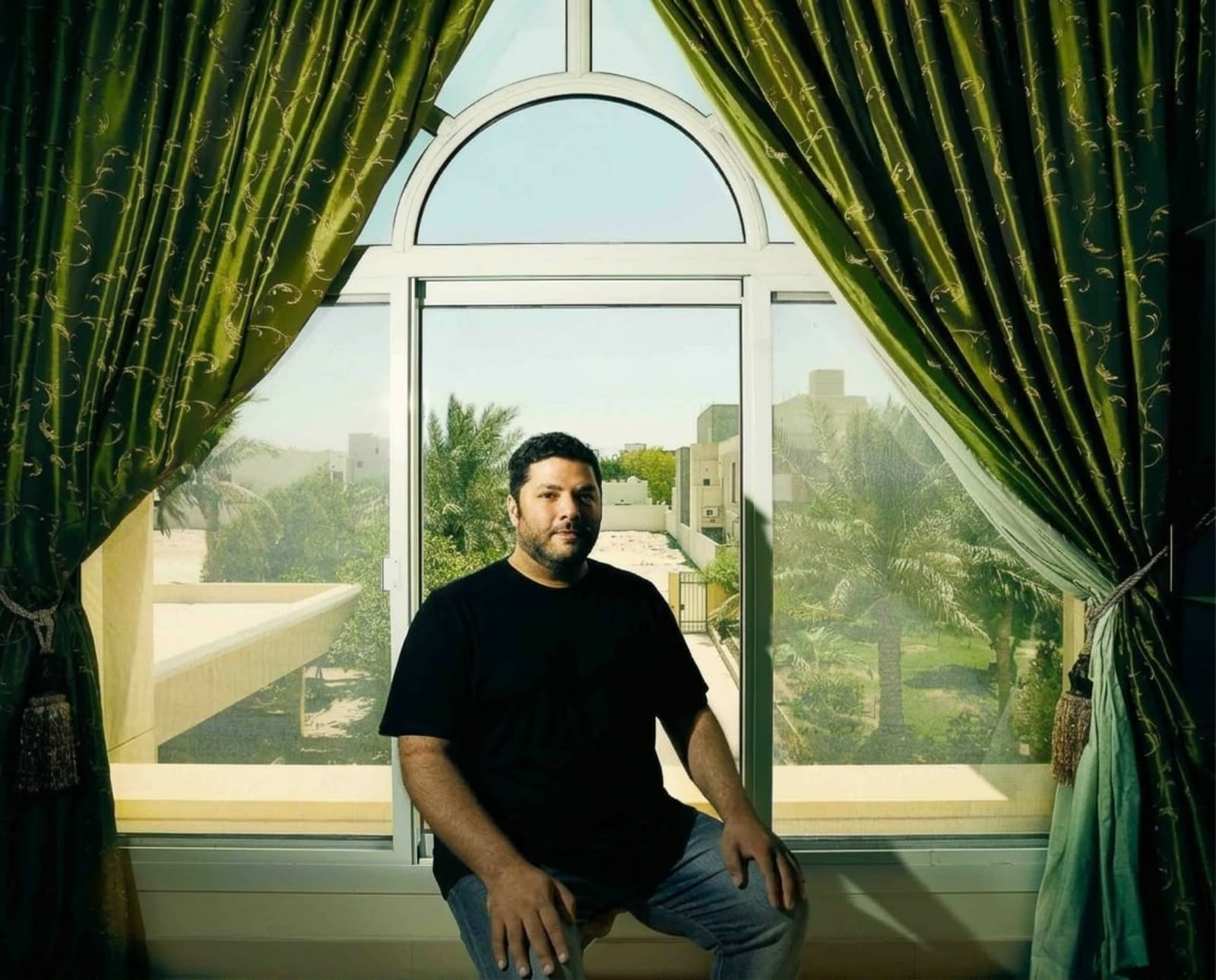 A portrait of Hasan Hujairi sitting in a window front, framed by green curtains